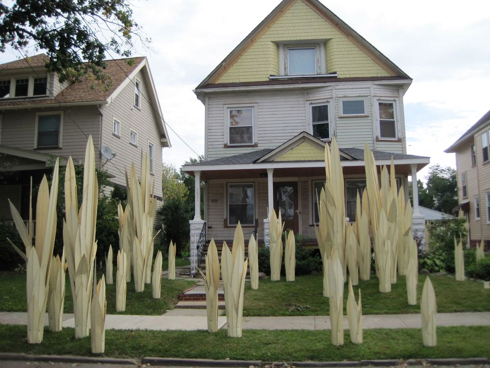 Tall pale sculptural blades planted across the front lawn of a wooden Cleveland house, with neighbouring houses either side.