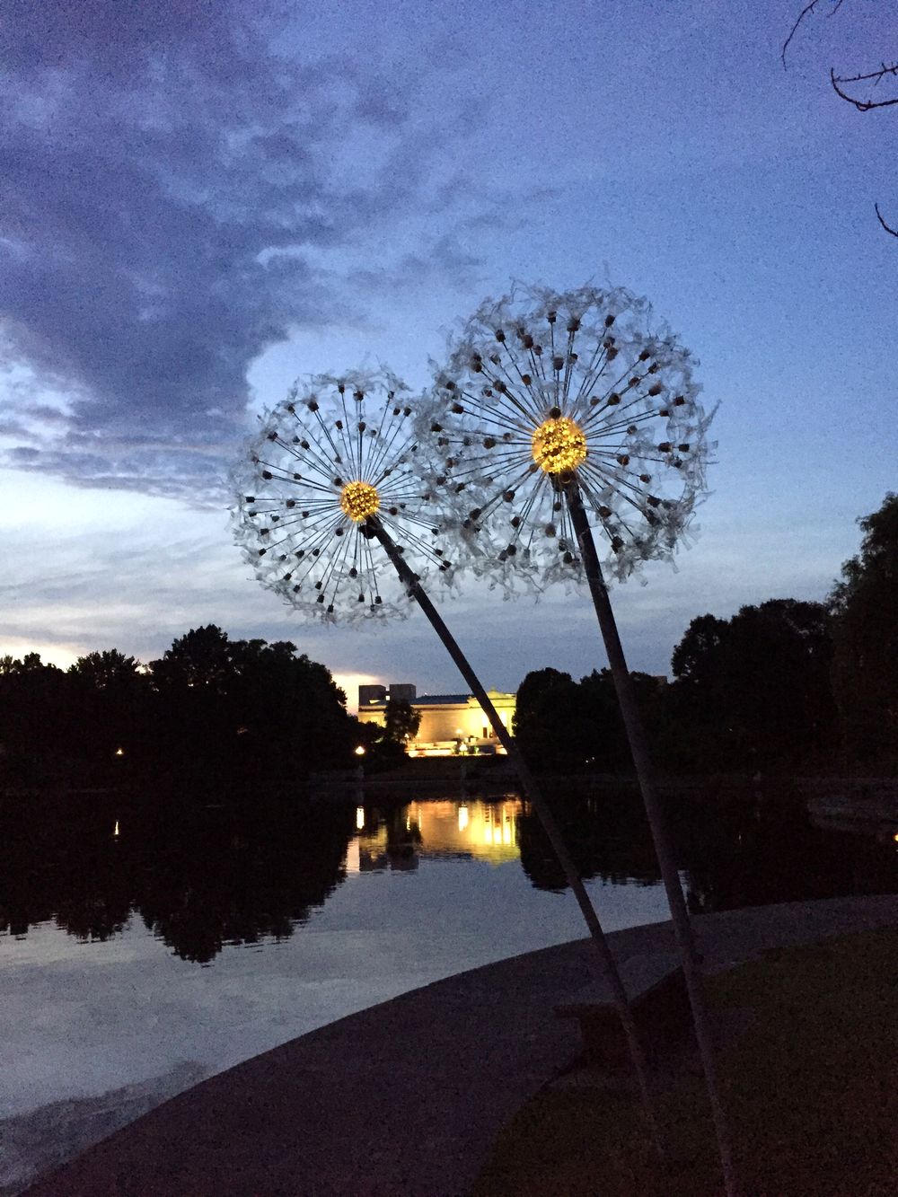 Two giant illuminated dandelion sculptures on tall stems at dusk, beside water, with the Cleveland skyline behind them.