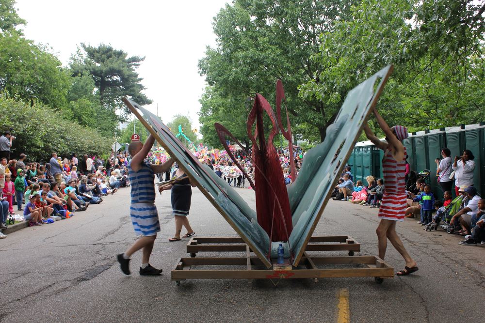 Two people in striped costumes wheeling a float carrying tall blue sculptural forms down a parade route.