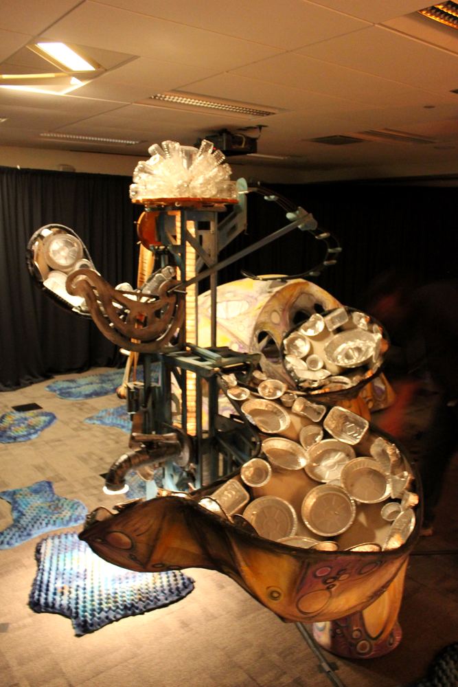 An assembled figure made from salvaged brass and tin vessels, standing on a patterned floor.