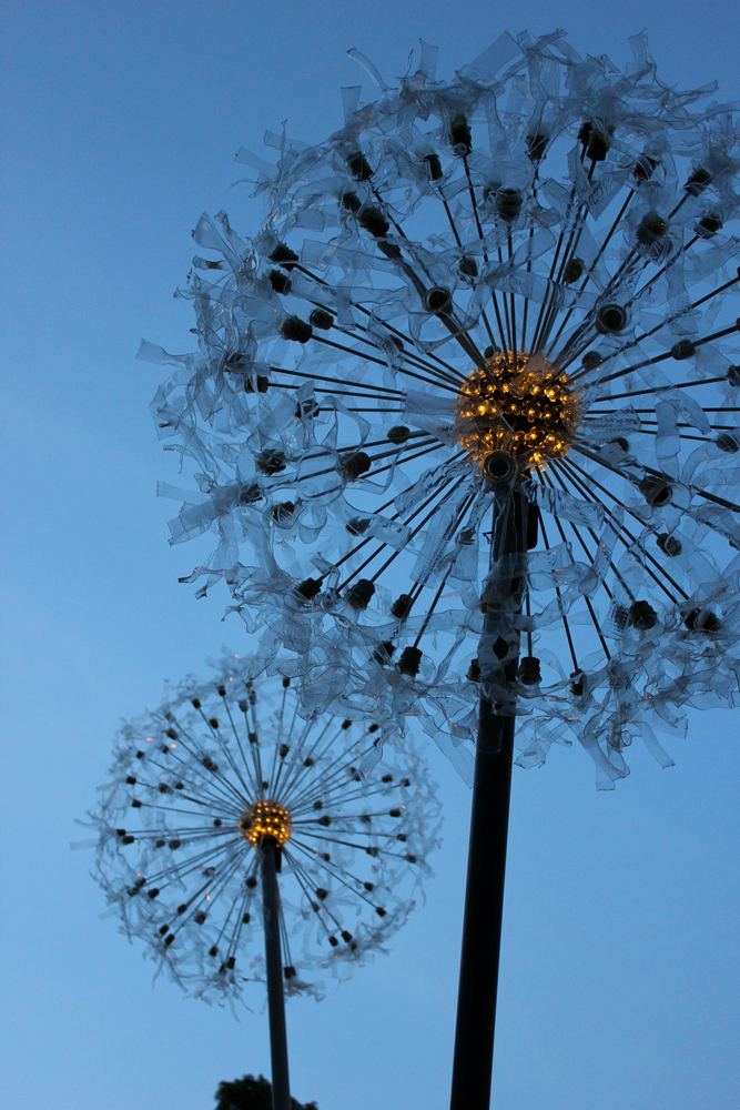 A single giant dandelion seedhead sculpture on a tall stem, photographed from below against a bright blue sky.