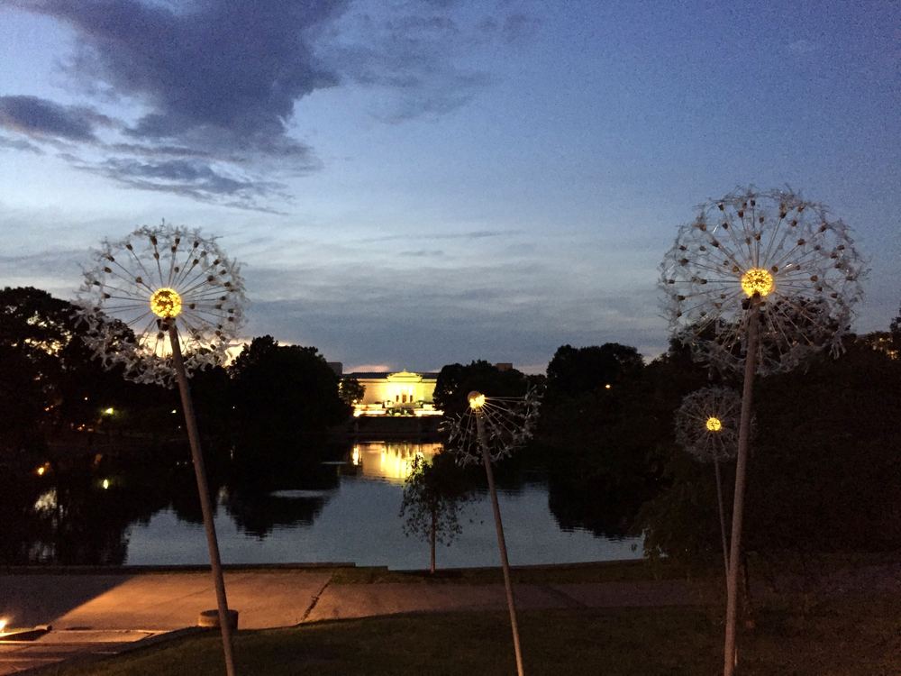 Two illuminated dandelion sculptures at twilight beside a lake, their lights reflected in the water.