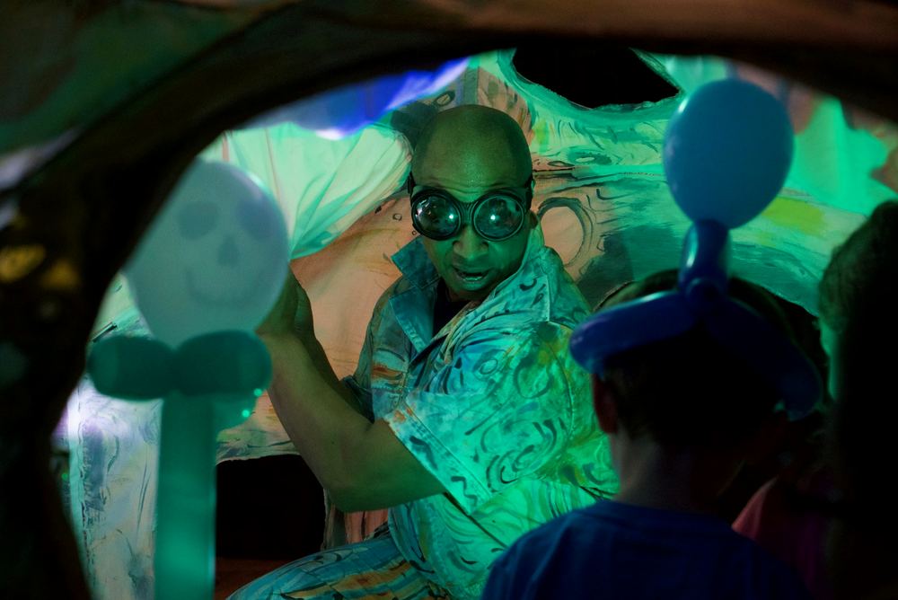 A performer lit in green inside a translucent sculptural creature, watched by a child in silhouette.