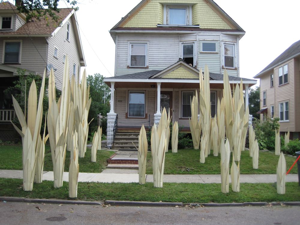 Tall pale sculptural blades planted across the front lawn of a wooden house on an ordinary street.