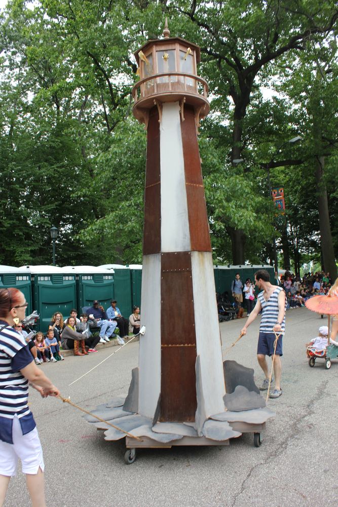 A tall lighthouse sculpture on wheels being walked along a parade route past a seated crowd.