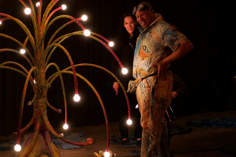 A sculptural tree hung with dozens of glowing bulbs on a darkened stage, two performers standing beneath it.