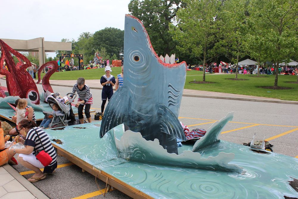 A blue shark sculpture cresting a wave on a parade float, with people gathered along the street behind it.