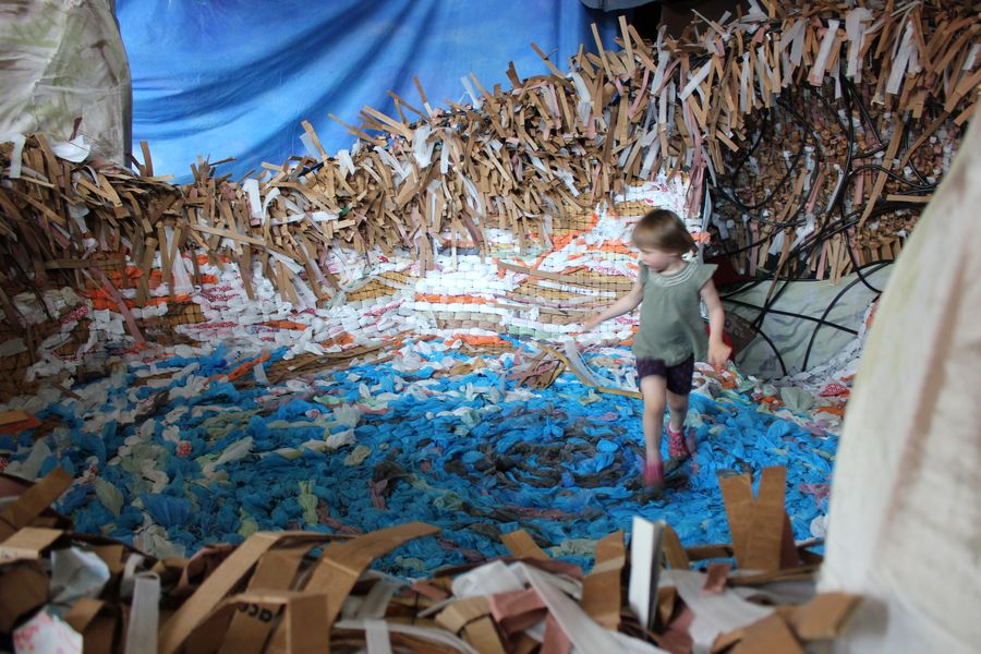 A child walking through a room-sized installation of painted cardboard waves and cut paper.
