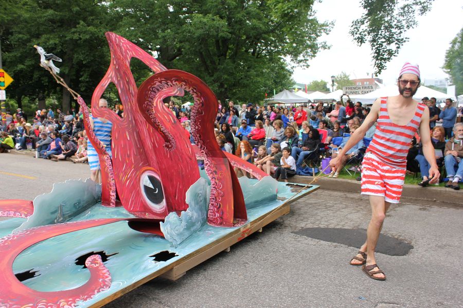 A large red squid sculpture riding a wheeled float down a street, a crowd seated along both kerbs.