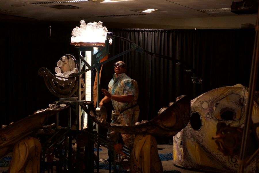 A performer on a darkened stage among large sculptural creature forms, one lit from within.