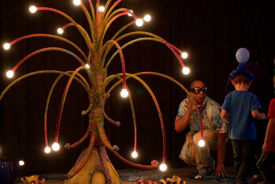 Ian crouching beside a tall sculptural tree strung with glowing bulbs, talking to children looking up at it.